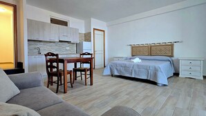 A compact hotel room with a kitchenette, a bed with a headboard, a bedside table, and a sofa. - Residenza Albachiara (Castelsardo)