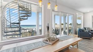 Un intérieur moderne agrémenté d’un escalier en colimaçon, de grandes fenêtres offrant une vue sur la plage et d’une table à manger en bois décorée de divers objets.