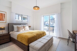 Ett modernt sovrum med en stor säng, ett sängbord, ett skrivbord och utsikt över staden genom stora fönster.