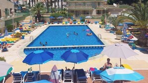 Μια πισίνα με πολλές διαδρομές, περιτριγυρισμένη από ξαπλώστρες και ομπρέλες, με φοίνικες και κτίρια στο βάθος.