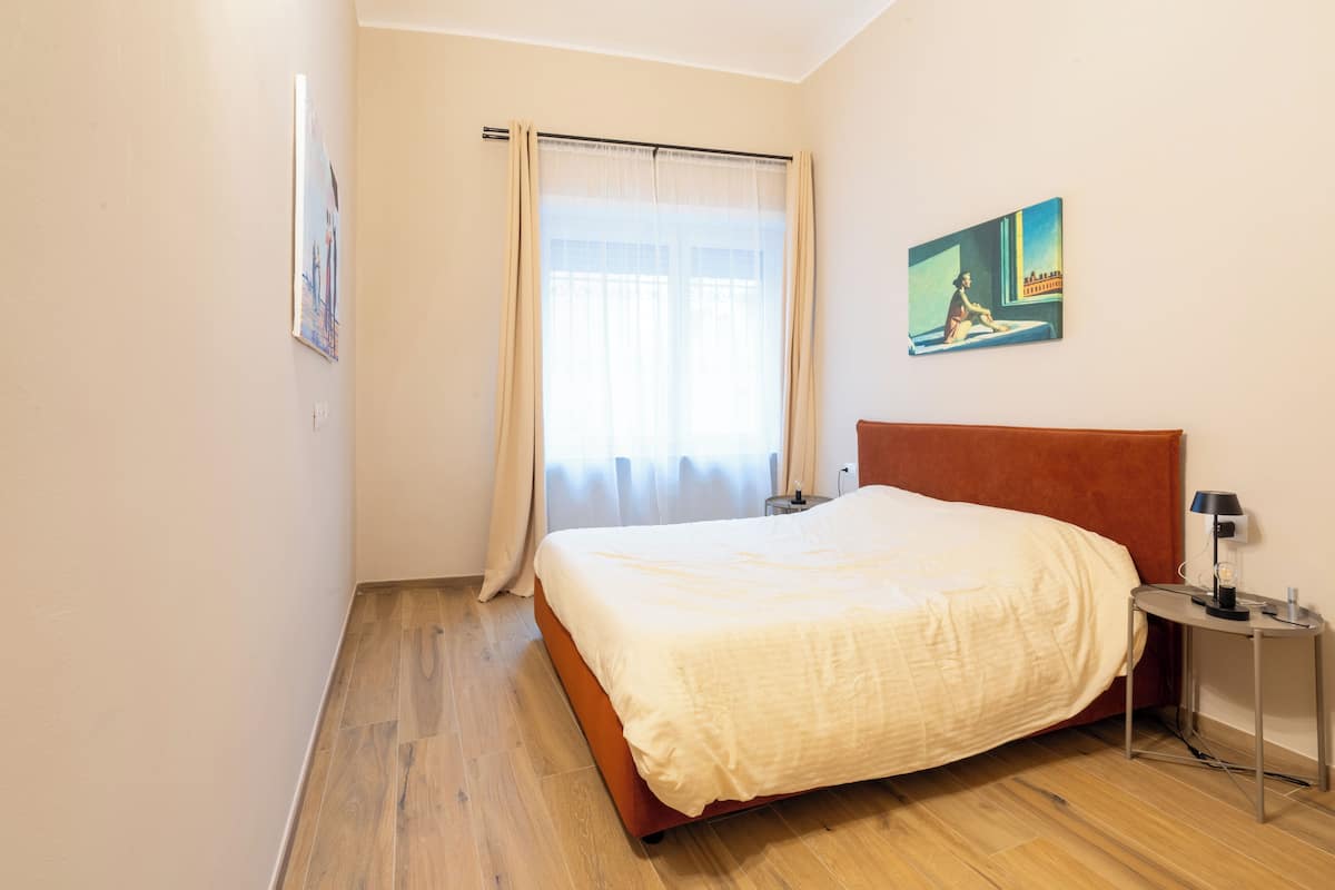 A bedroom with a bed, a nightstand, a painting on the wall, and a window with curtains.