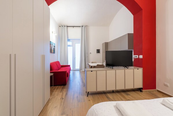A modern hotel room with a wooden floor, a bed, a red sofa, a TV, and a cabinet.