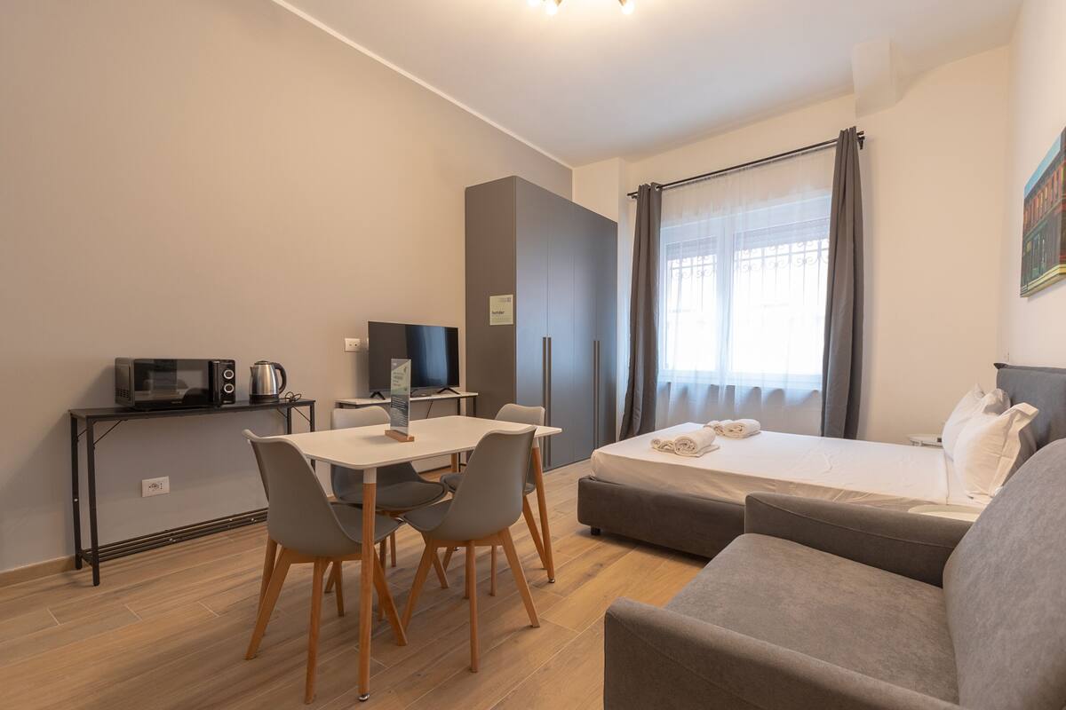 A compact hotel room with a bed, a sofa, a dining table with chairs, a TV, and a microwave.