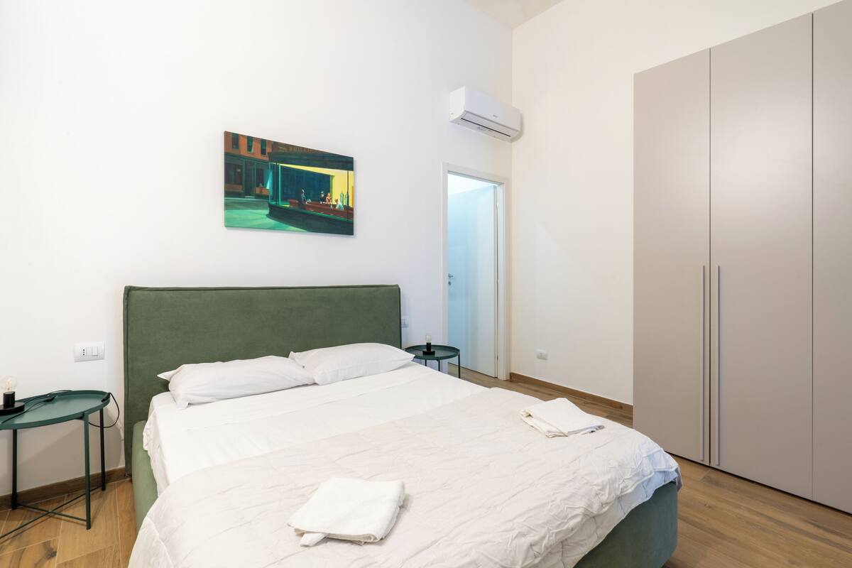A hotel room with a large bed, a green headboard, a painting on the wall, a wardrobe, and an air conditioning unit.