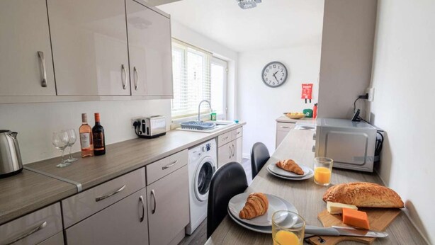 Cocina funcional con lavadora, tostadora y microondas. Se observa una mesa de comedor con un croissant, queso y jugo.