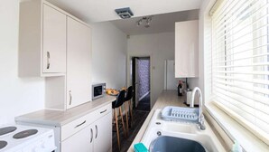 Cocina compacta con armarios blancos, estufa, lavamanos y ventana con persianas.