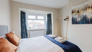 A bedroom with a bed, curtains, a radiator, a painting, and a window with a view of houses. - Fabulous 3 bedroom Home in Redcar (Redcar)