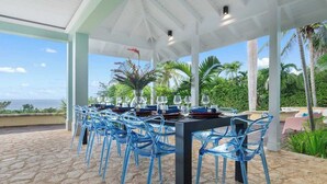 An outdoor dining area with blue chairs and a table set for a meal, overlooking a scenic view of the ocean and palm trees. - Greens All (Montego Bay)