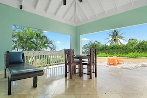 A covered patio with a wooden bench, a table with chairs, and a view of palm trees and greenery.