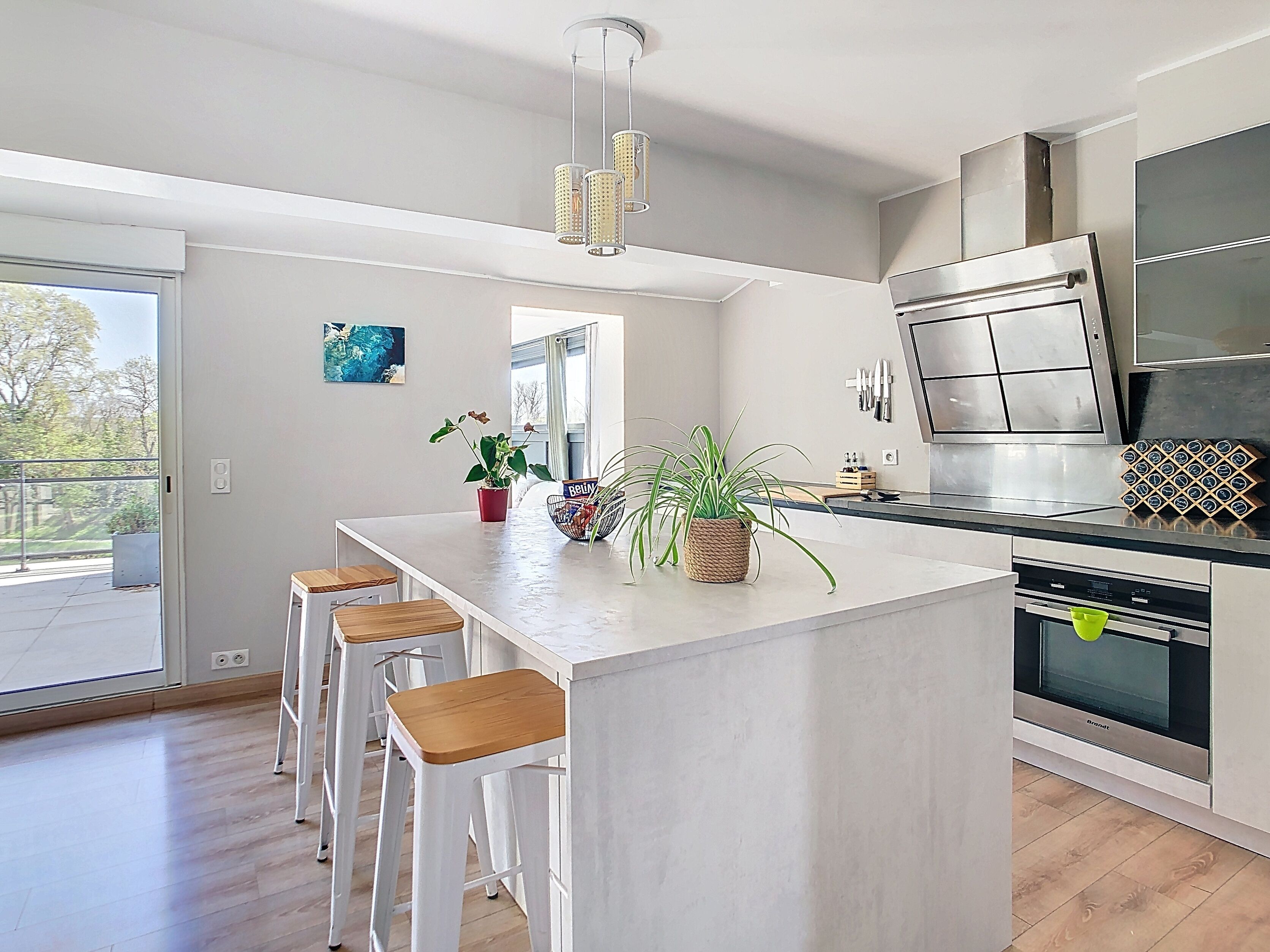 Une cuisine moderne dotée d’un îlot central, de tabourets de bar, d’appareils électroménagers en acier inoxydable et offrant une vue sur l’extérieur.