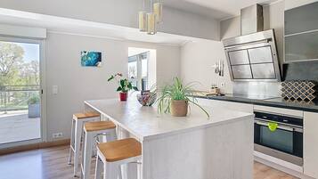 Une cuisine moderne dotée d’un îlot central, de tabourets de bar, d’appareils électroménagers en acier inoxydable et offrant une vue sur l’extérieur.