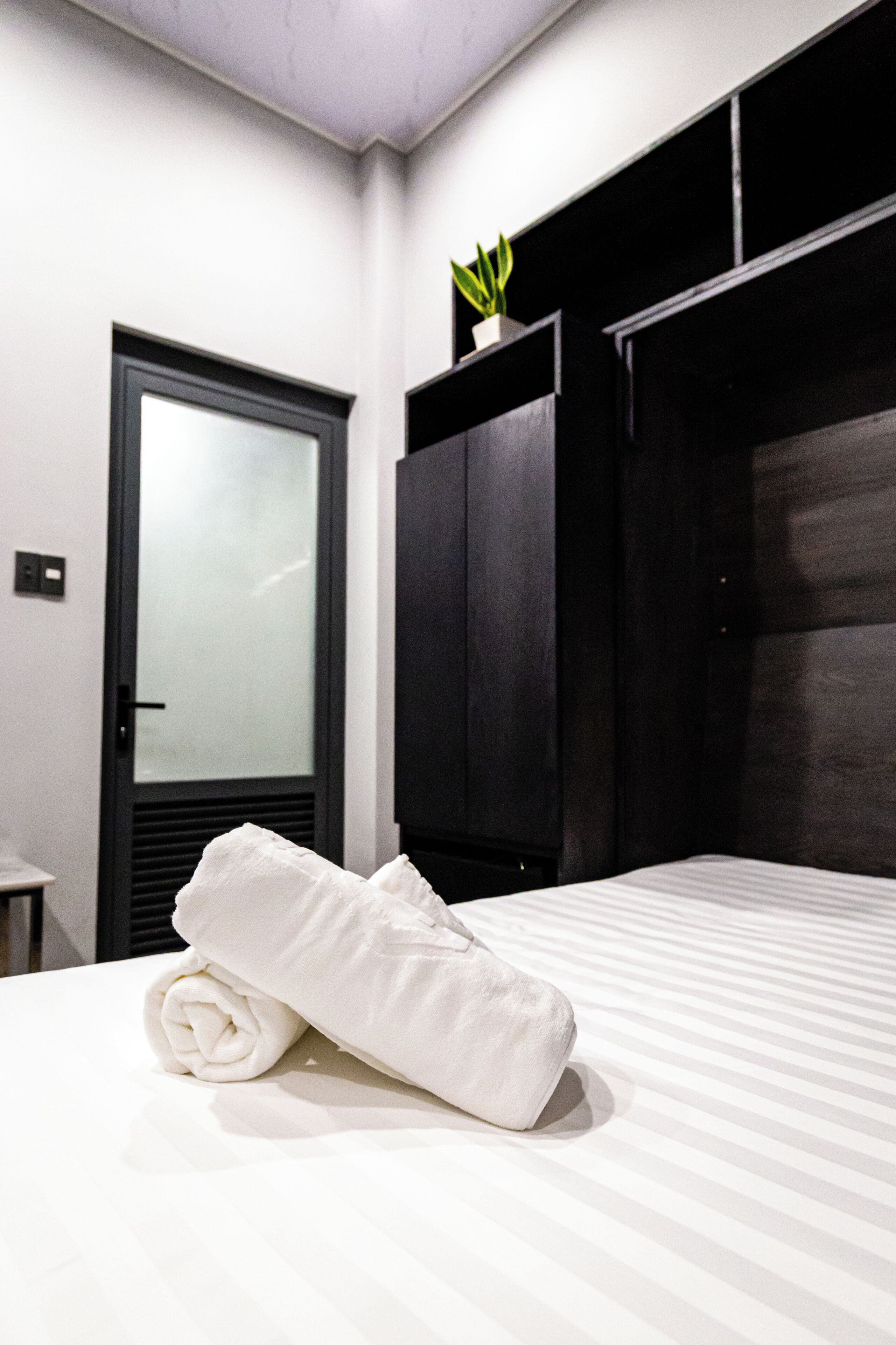 A hotel room with a bed, a dark wooden wardrobe, a white towel on the bed, and a small potted plant on the wardrobe.