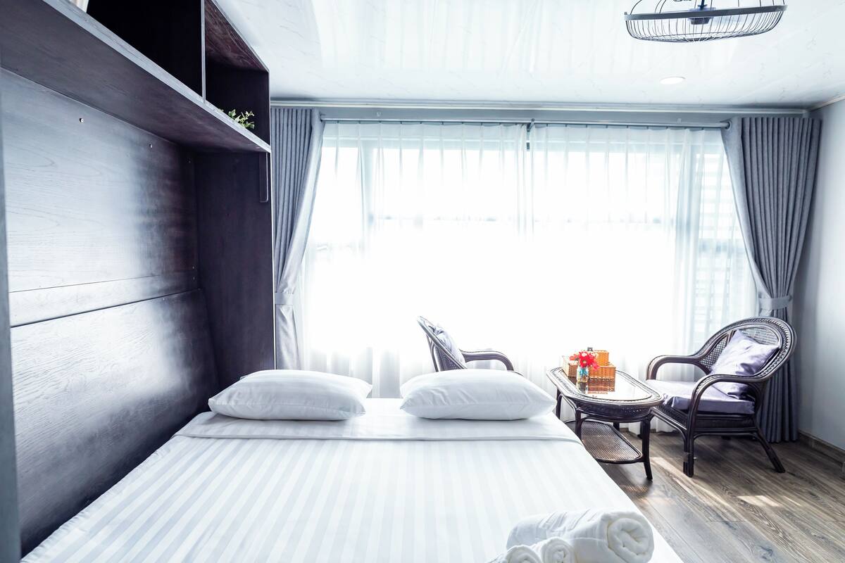 Een hotelkamer met een bed, twee stoelen, een tafeltje met een vaas en een raam met gordijnen.
