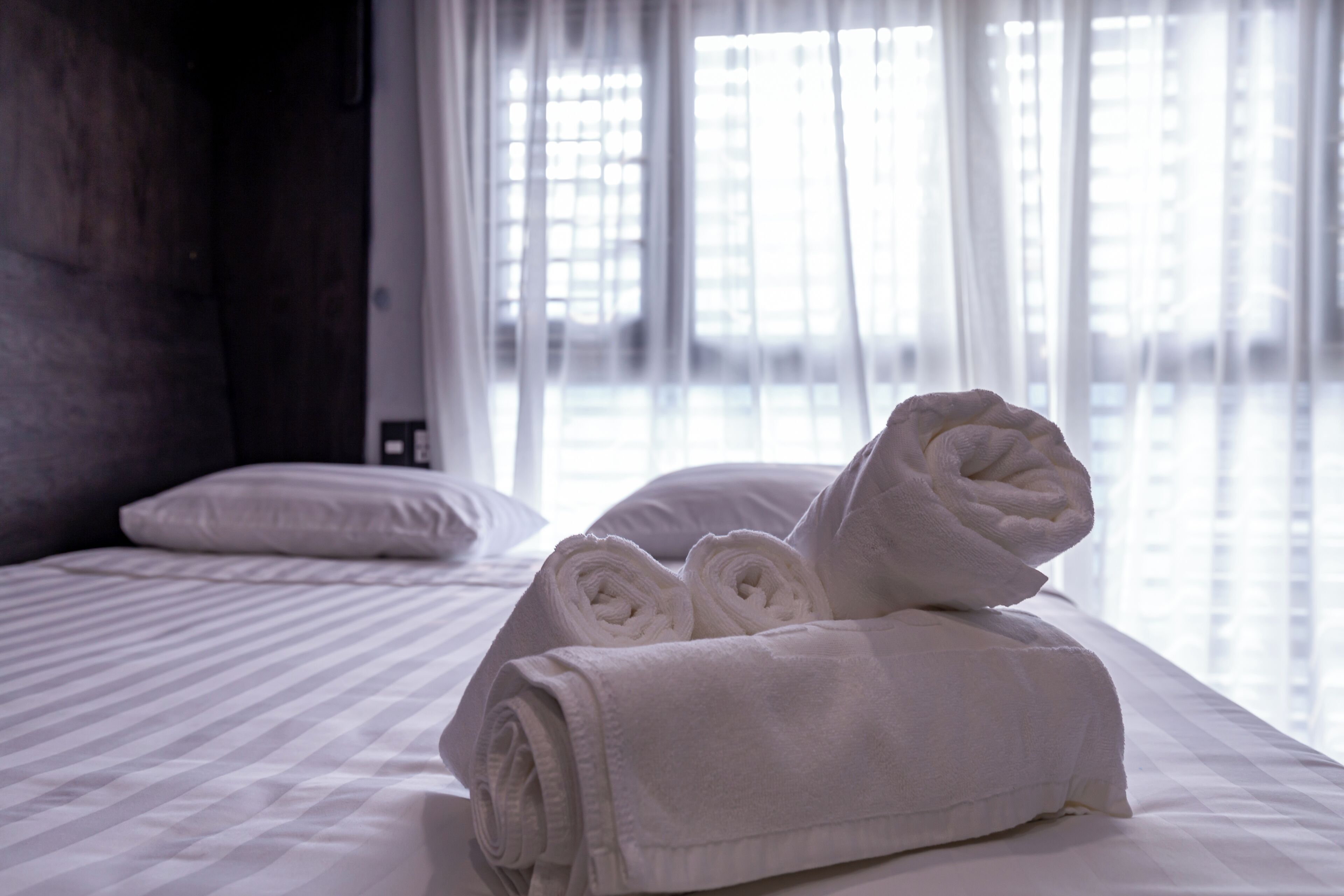 A neatly made bed with rolled white towels and a pillow, a window with sheer curtains, and a plain wall.