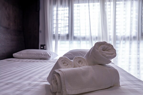 A neatly made bed with rolled white towels and a pillow, a window with sheer curtains, and a plain wall.