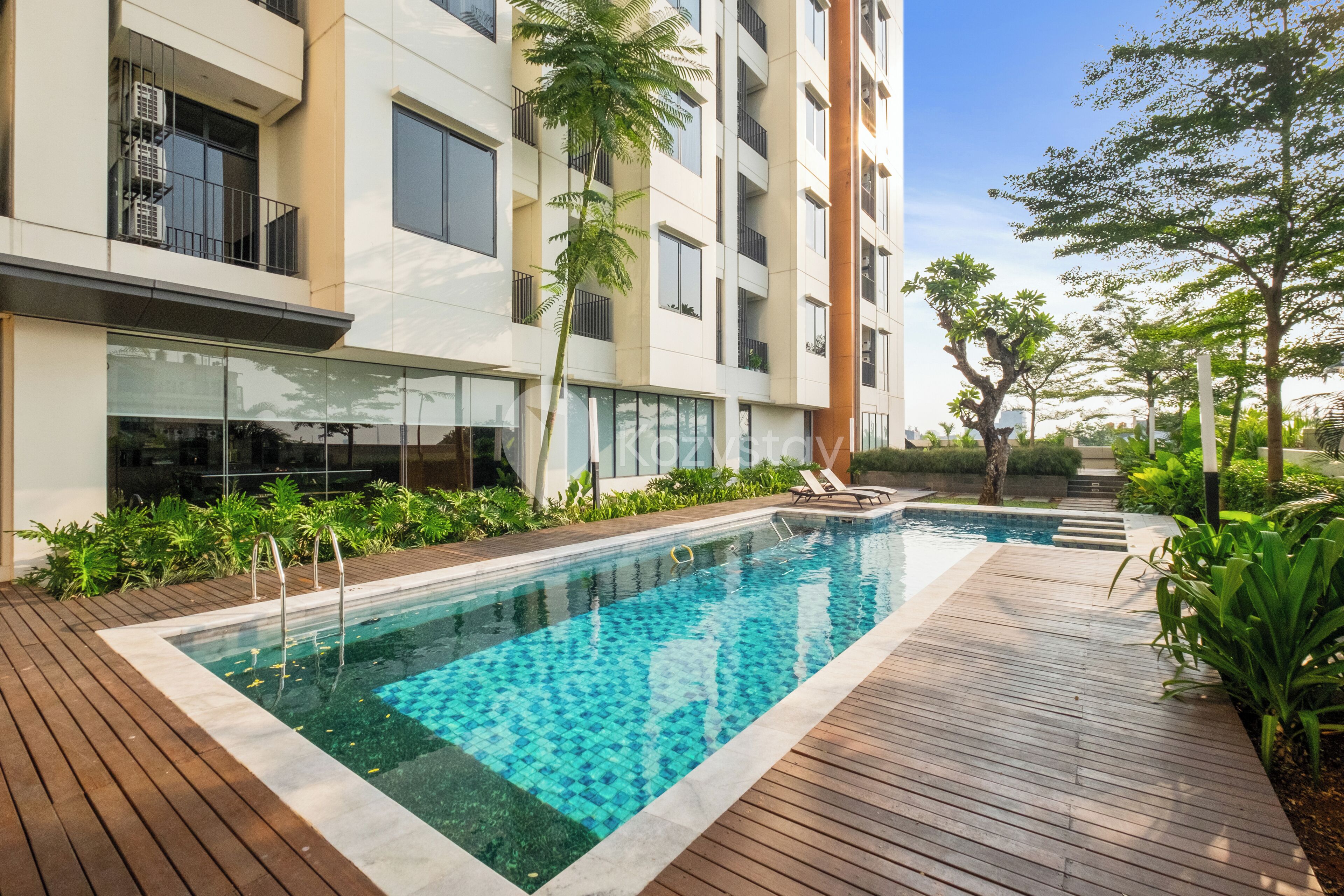 Un edificio moderno con una piscina rectangular rodeada de terrazas de madera y abundante vegetación.