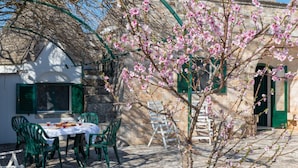 A table set outdoors with chairs, a stone building with green shutters, and a flowering tree.
