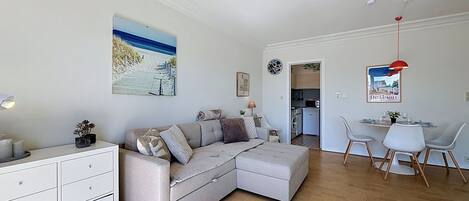 Una sala de estar moderna con un sofá, un comedor con sillas, una cocina al fondo y un cuadro con motivos playeros en la pared.