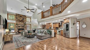 A spacious living area with a stone fireplace, a wooden staircase, and a kitchen with modern appliances.