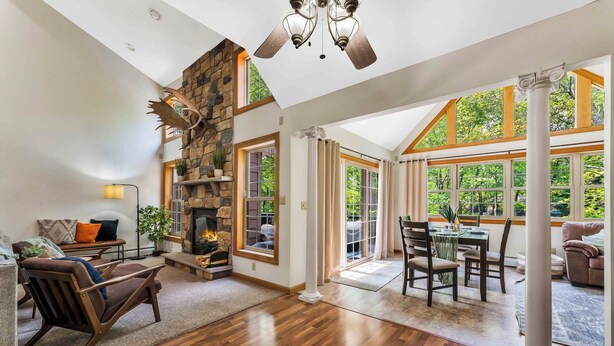 A spacious living room with a stone fireplace, wooden flooring, and large windows offering a view of the outdoors.
