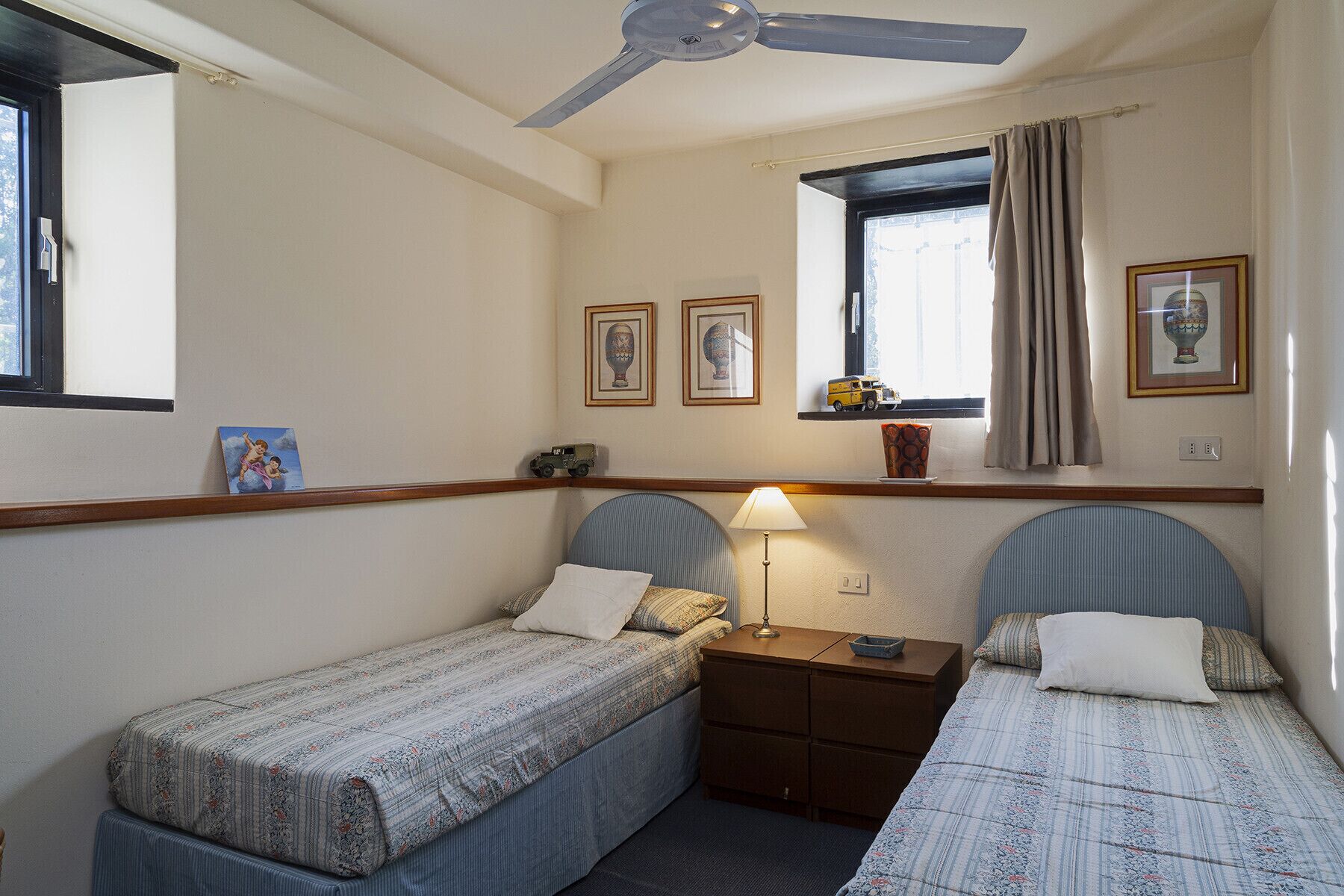 A small, cozy room with two single beds, a nightstand, a lamp, and a ceiling fan.