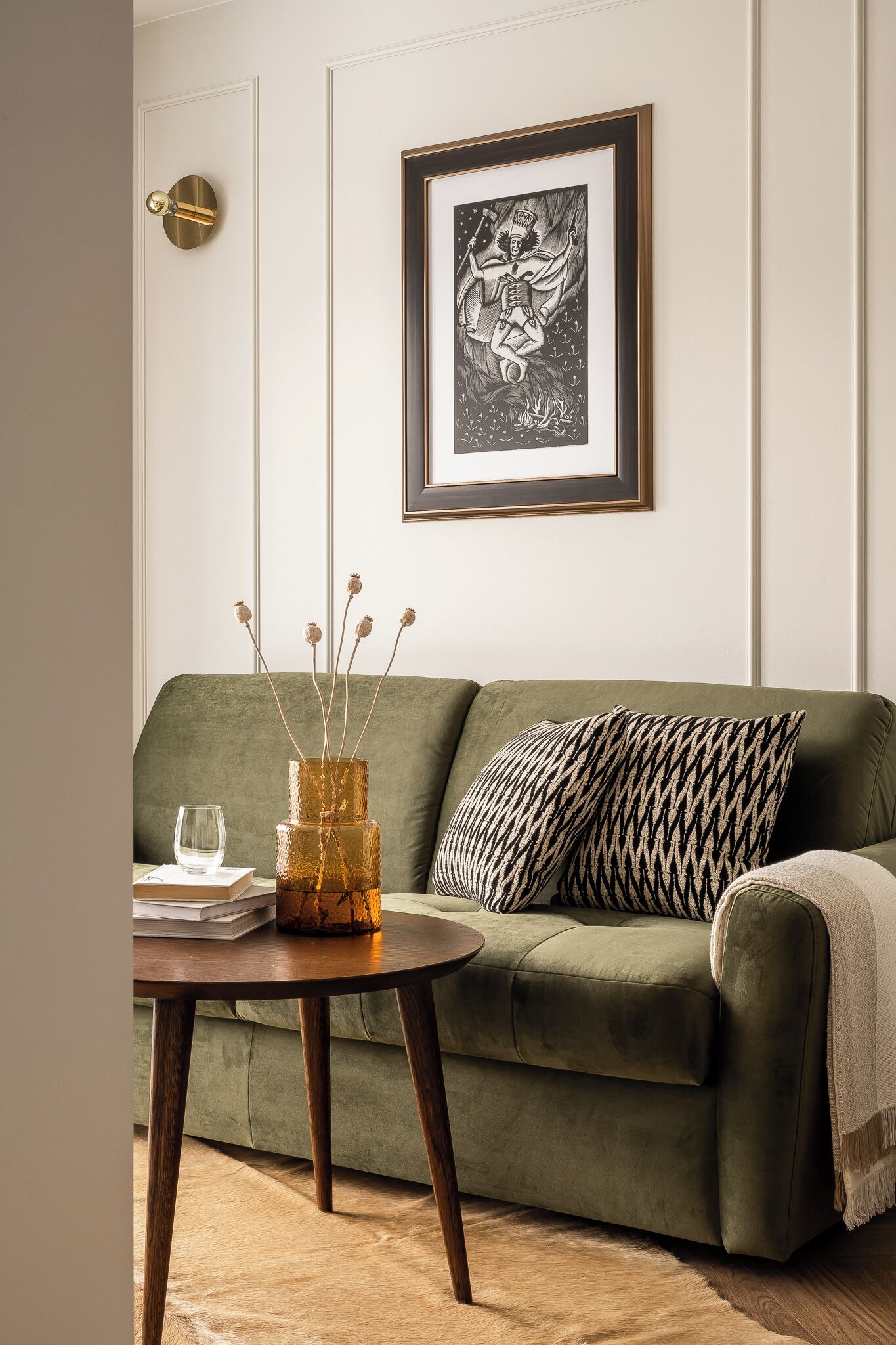 A green sofa with a patterned pillow, a framed artwork above it, and a table with a vase and books.