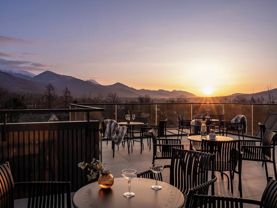 An outdoor dining area with tables and chairs, overlooking a mountainous landscape during sunset.