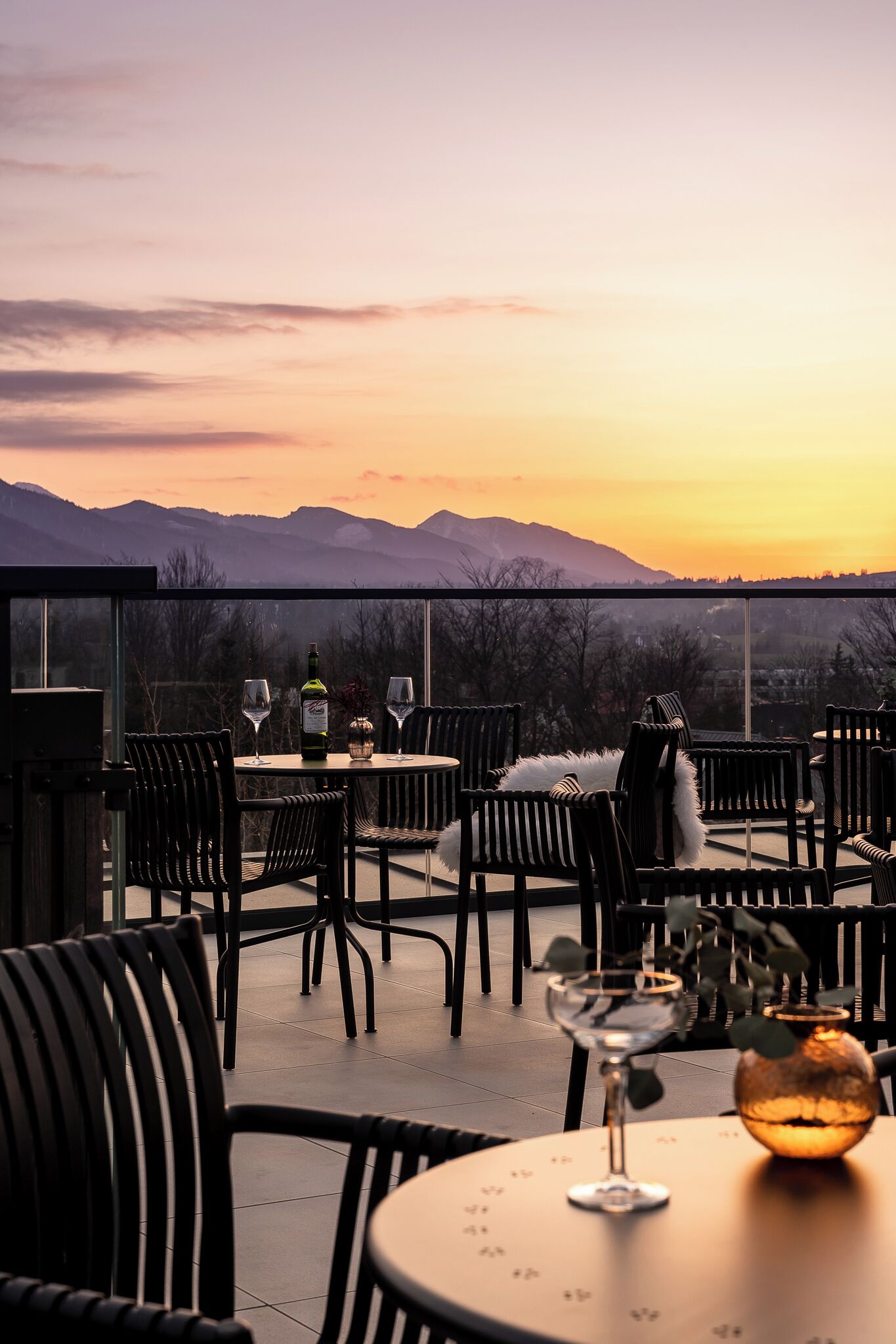 A terrace with a table set for two, featuring a bottle of wine and two glasses, overlooking a mountainous landscape during sunset.