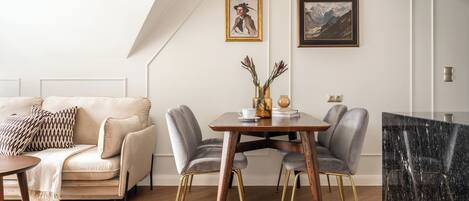 A modern living room with a sofa, dining table, and chairs. There are two framed pictures on the wall, a vase with flowers on the table, and an air conditioning unit above the table.