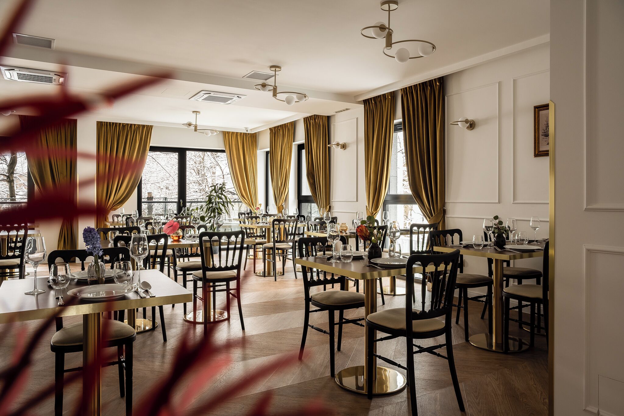 Een ruime eetruimte met meerdere tafels en stoelen, grote ramen voorzien van gordijnen en moderne verlichtingsarmaturen.