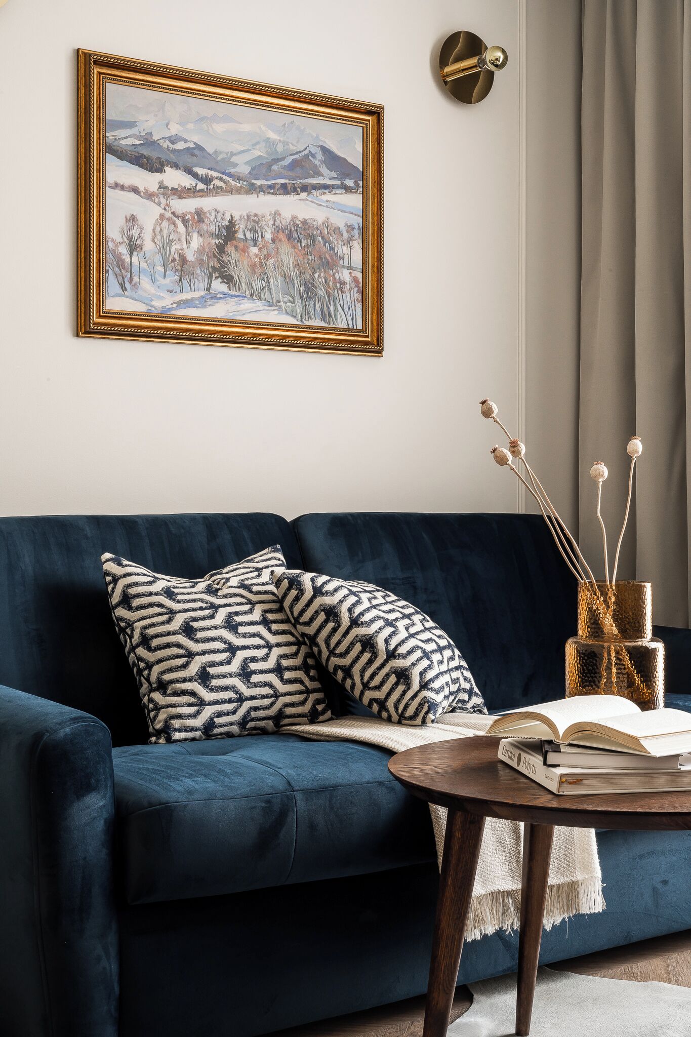 A dark blue sofa with patterned cushions, a framed landscape painting, a small round table with a book, and a decorative vase with branches.