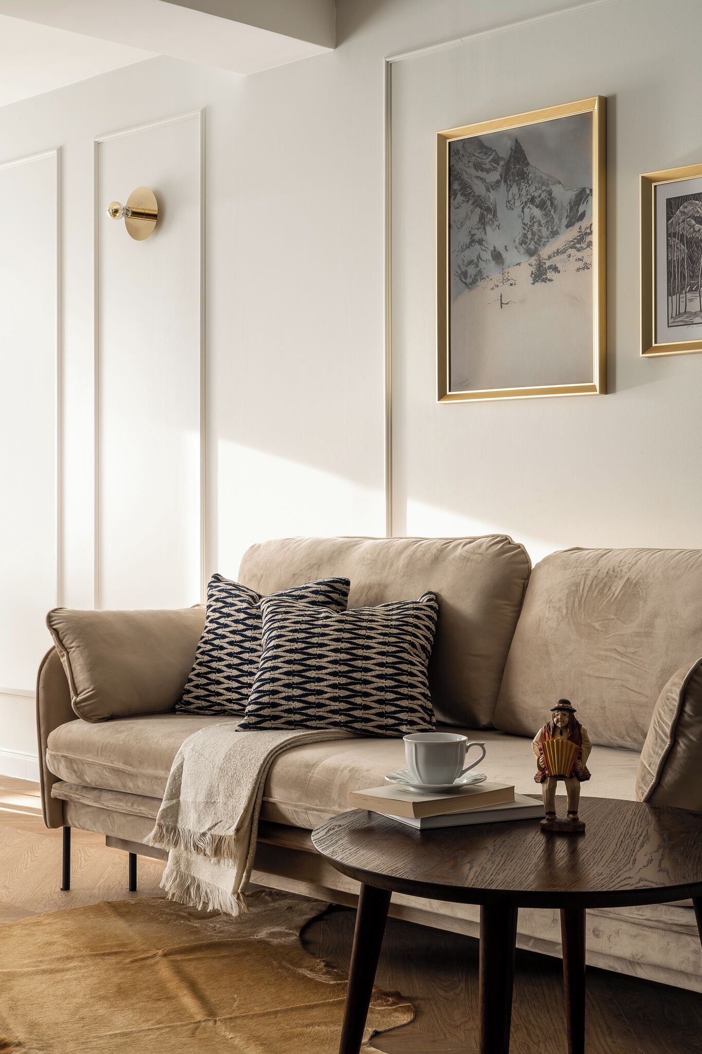 A modern living room with a beige sofa, patterned cushions, a small table with a cup and book, and framed artwork on the wall.