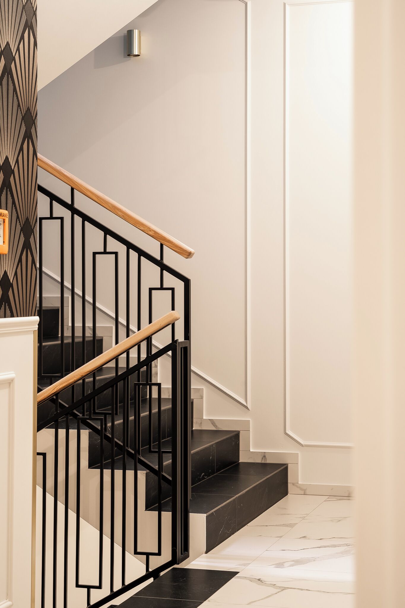 A modern staircase with black railings and wooden handrails, leading to a landing with a light-colored wall and a geometric-patterned wall on the left.