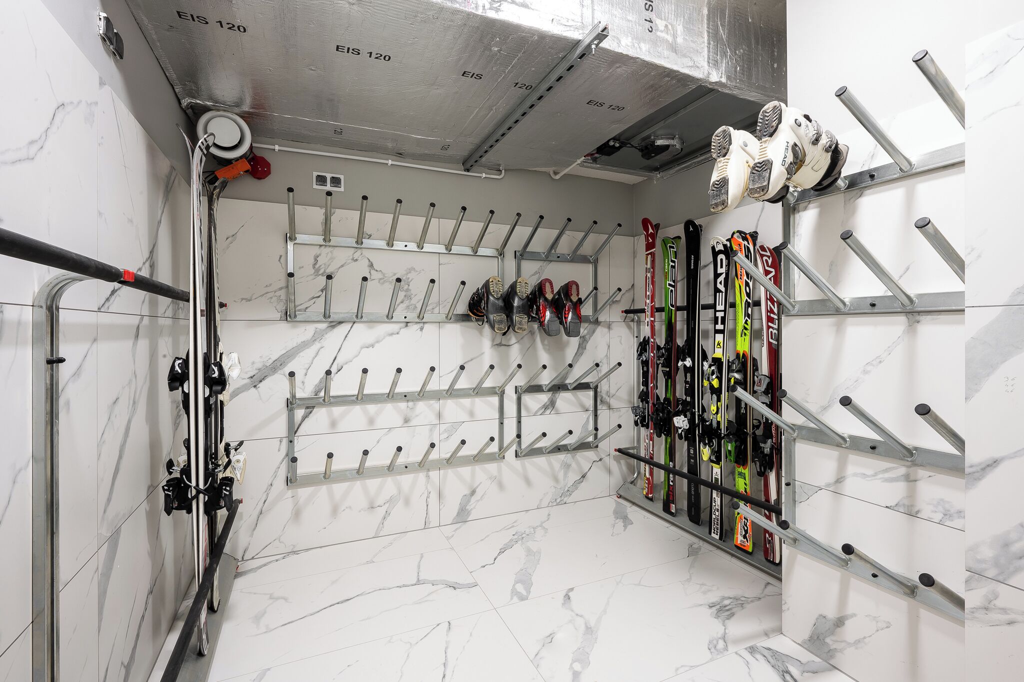 A ski storage area with multiple racks for skis and snowboards, a metal ceiling, and marble flooring.
