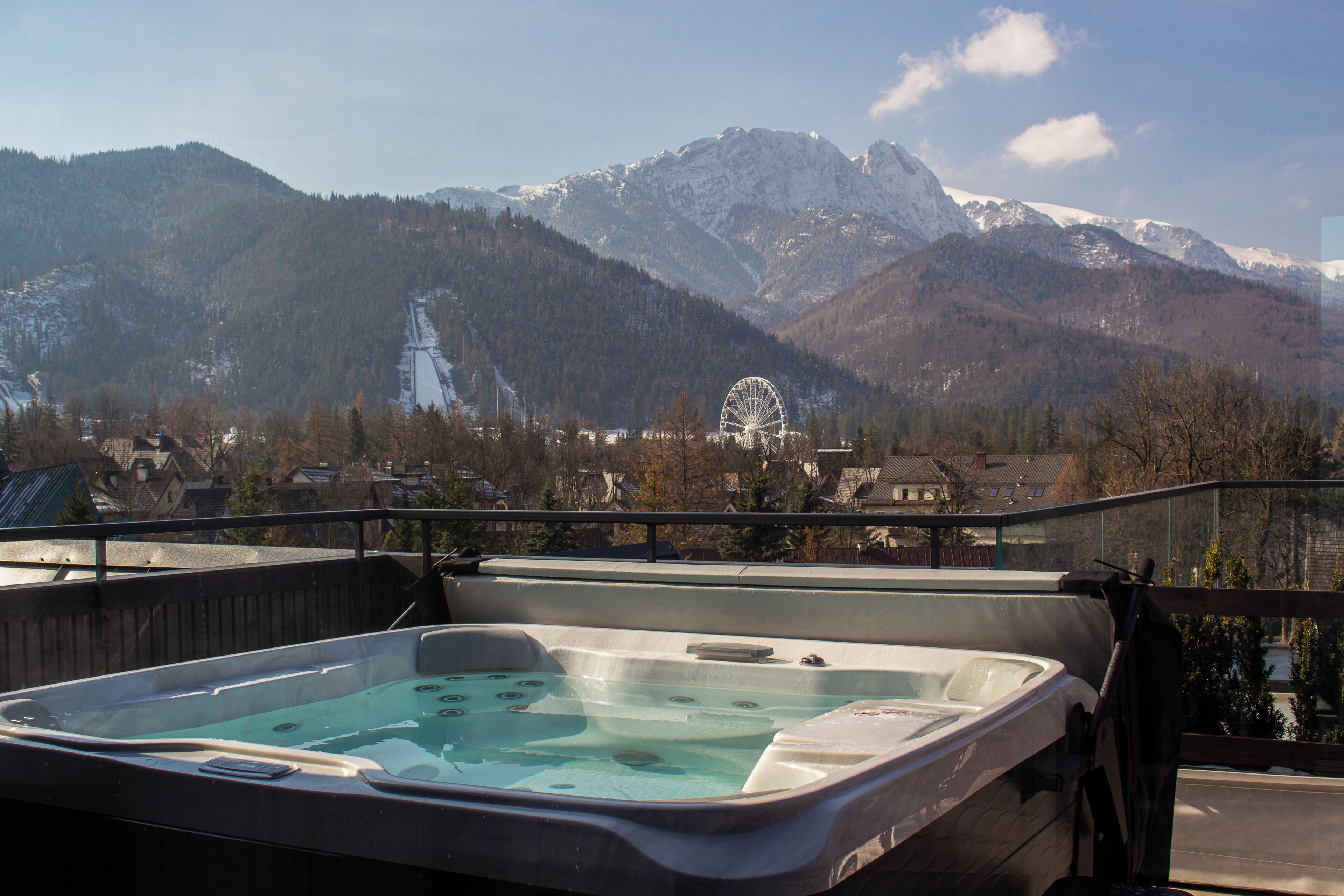 Un jacuzzi en un balcón con vistas a la montaña, una noria y casas al fondo.