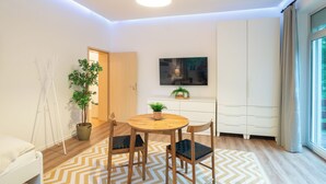 Ein modernes Wohnzimmer mit einem runden Holztisch, zwei Stühlen, einer Zimmerpflanze, einem an der Wand befestigten Fernseher und einem großen Fenster mit Vorhängen.