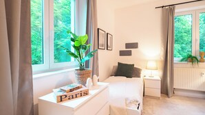 Ein Schlafzimmer mit Bett, Nachttisch mit Lampe, Fenster mit Vorhängen und Blick ins Grüne.