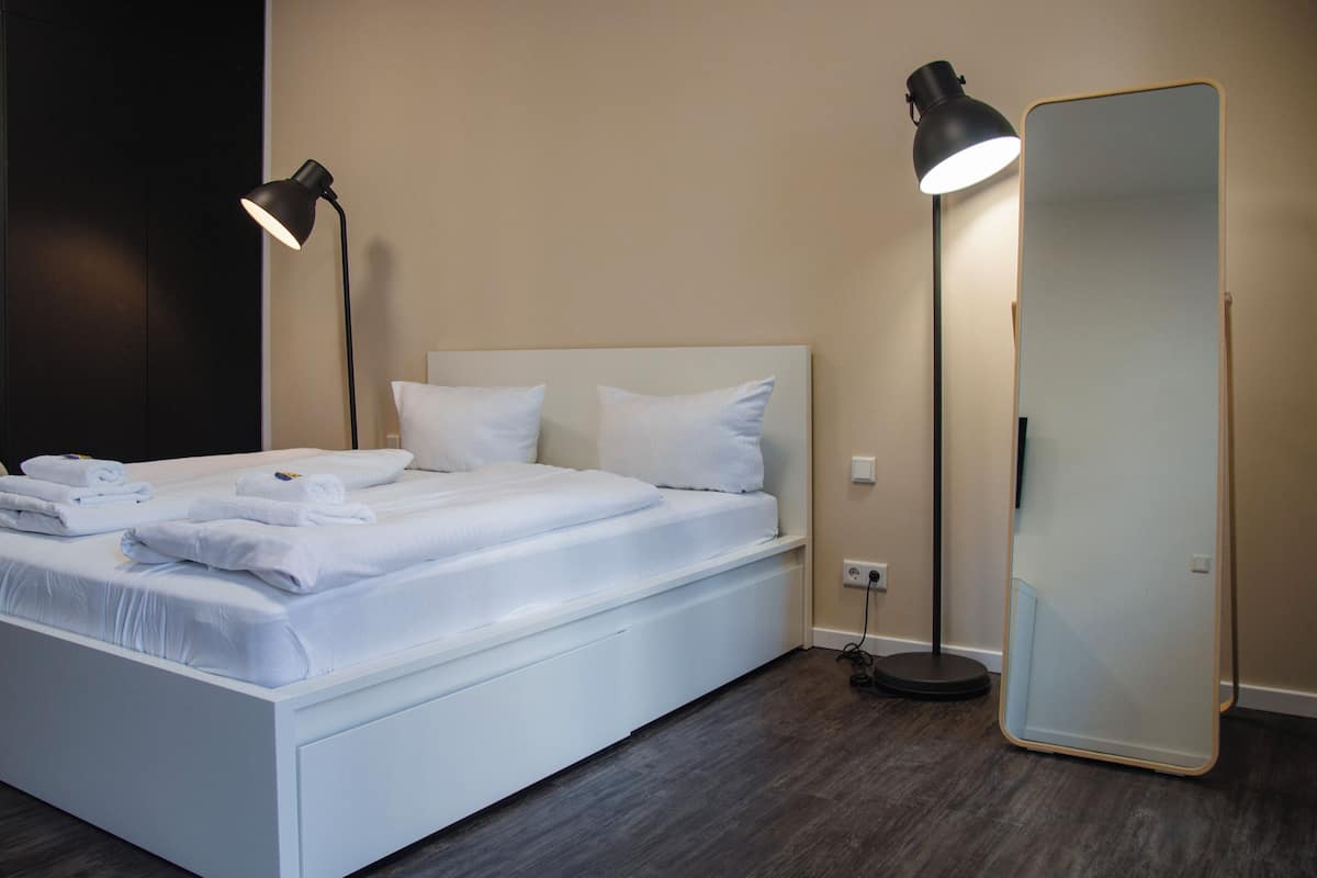 A neatly made bed with white linens and towels, a large mirror, and two floor lamps in a modern room with dark wooden flooring.