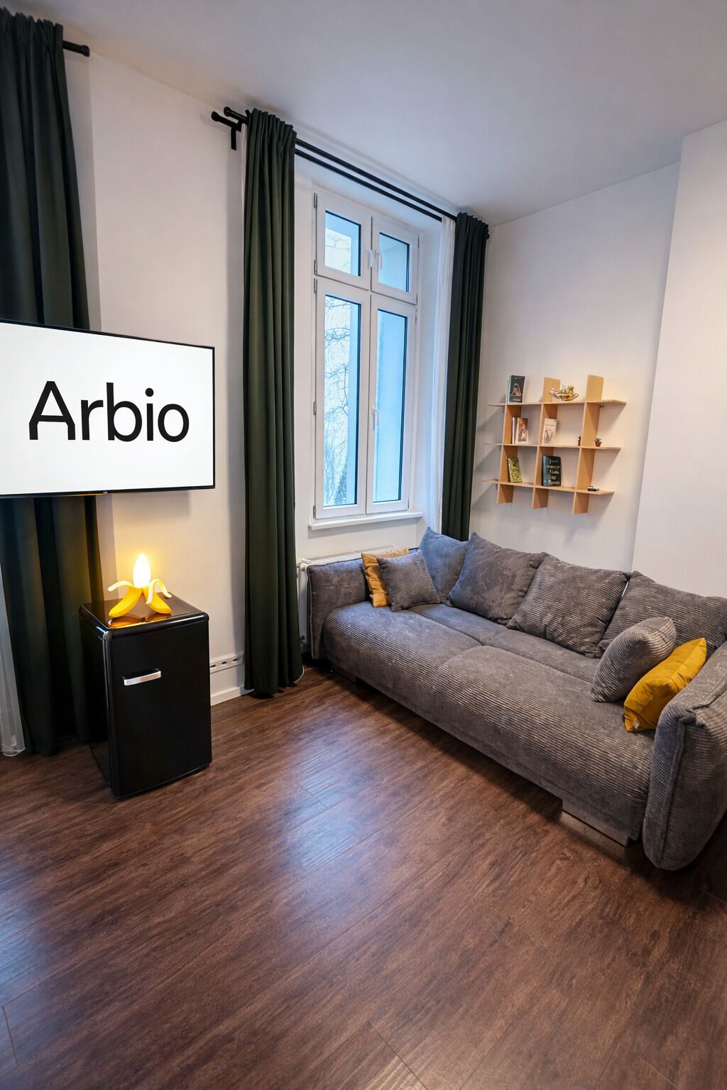 Een moderne woonkamer met een bank, een klein zwart kastje, een raam met gordijnen en een plank met boeken en decoratieve objecten.