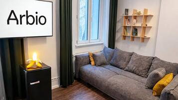 Een moderne woonkamer met een bank, een klein zwart kastje, een raam met gordijnen en een plank met boeken en decoratieve objecten.