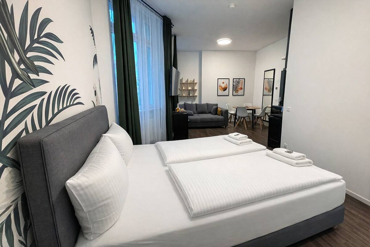 A modern hotel room with a large bed, a sofa, a dining table, and a view of the city.