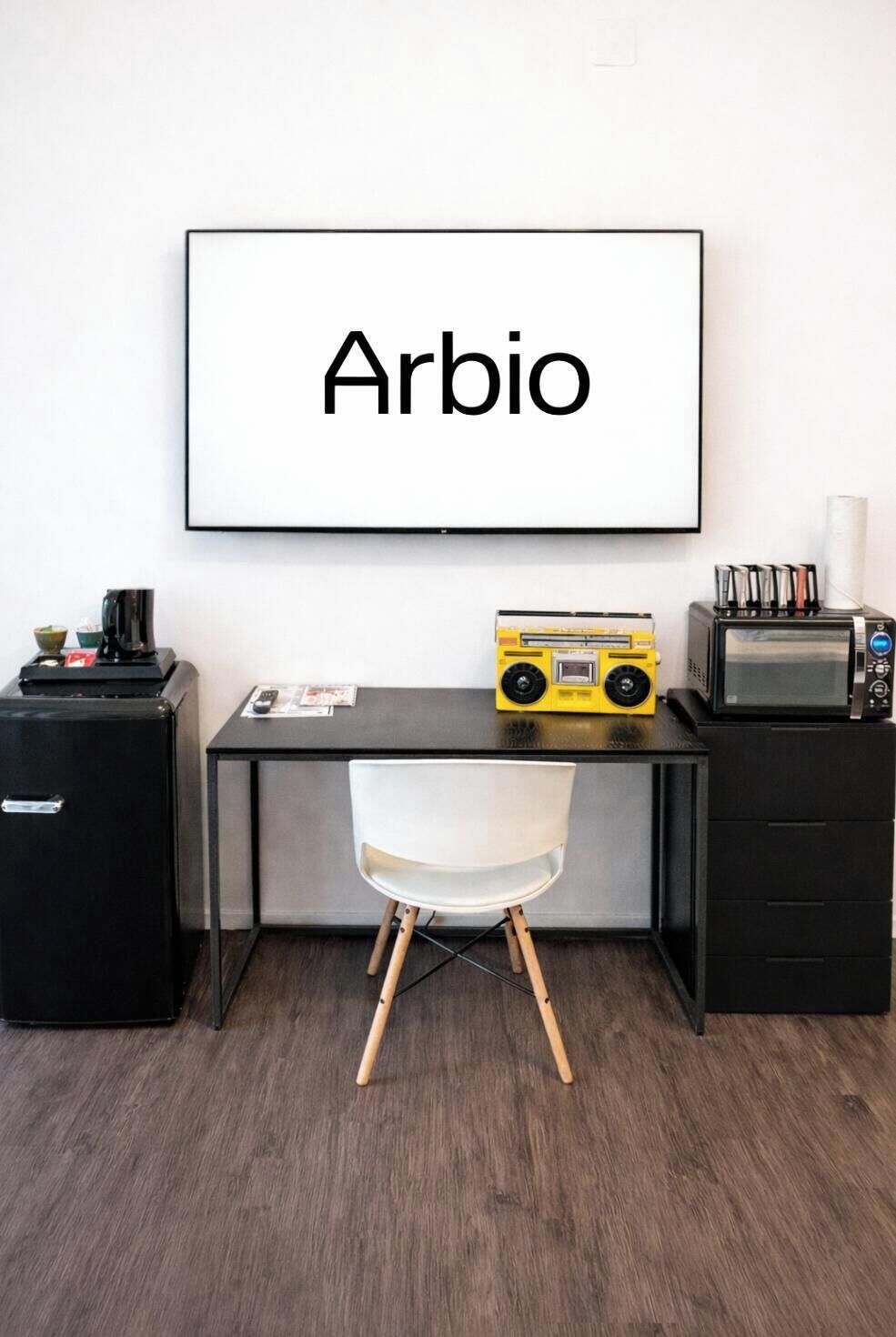 A modern room with a desk, a chair, a radio, a coffee machine, and a framed word 'Arbio' on the wall.