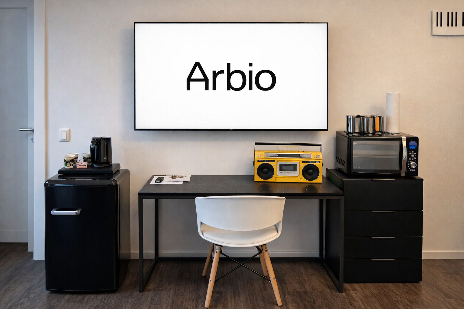 A compact room with a desk, a chair, a coffee machine, a radio, a microwave, and a small refrigerator.
