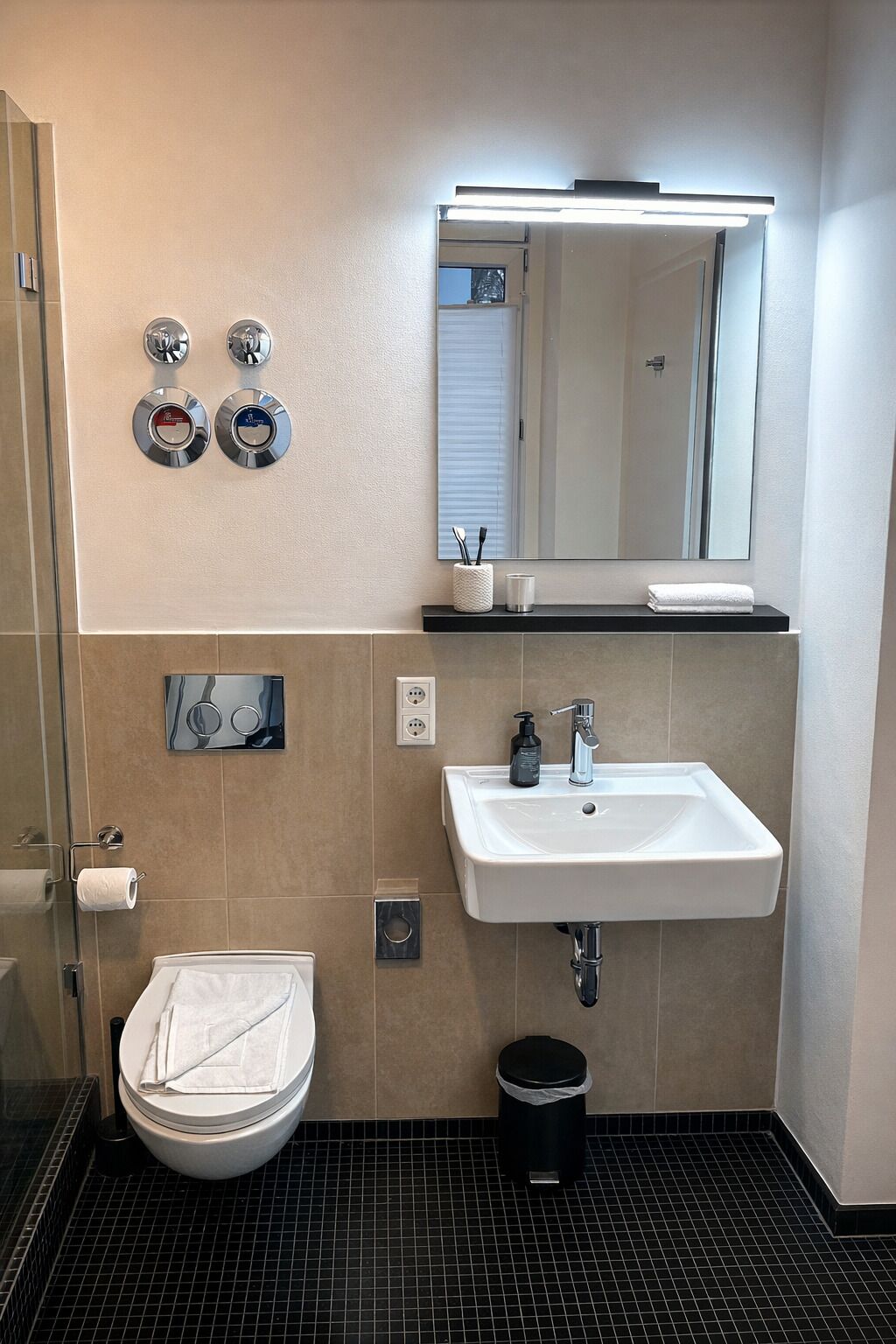 A bathroom with a white sink, mirror, and toilet. There are two towel dispensers, a soap dispenser, and a trash can.