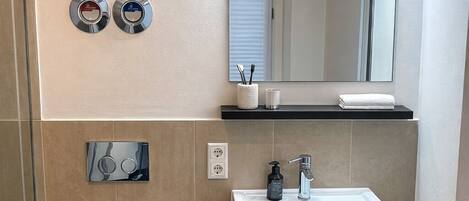 A bathroom with a white sink, mirror, and toilet. There are two towel dispensers, a soap dispenser, and a trash can.