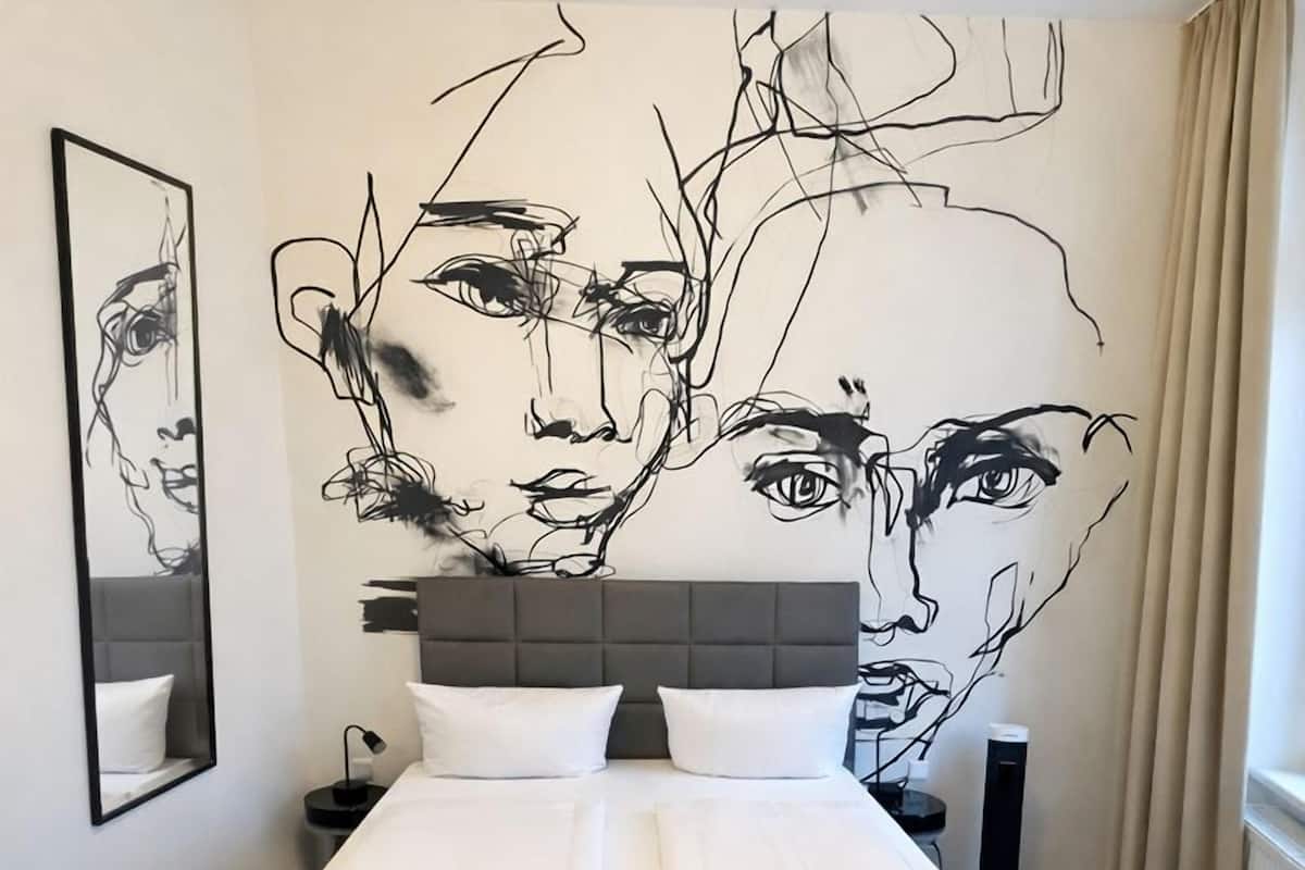 A hotel room with a large bed, a headboard, a wall-mounted artwork, a bedside table, and a window with curtains.