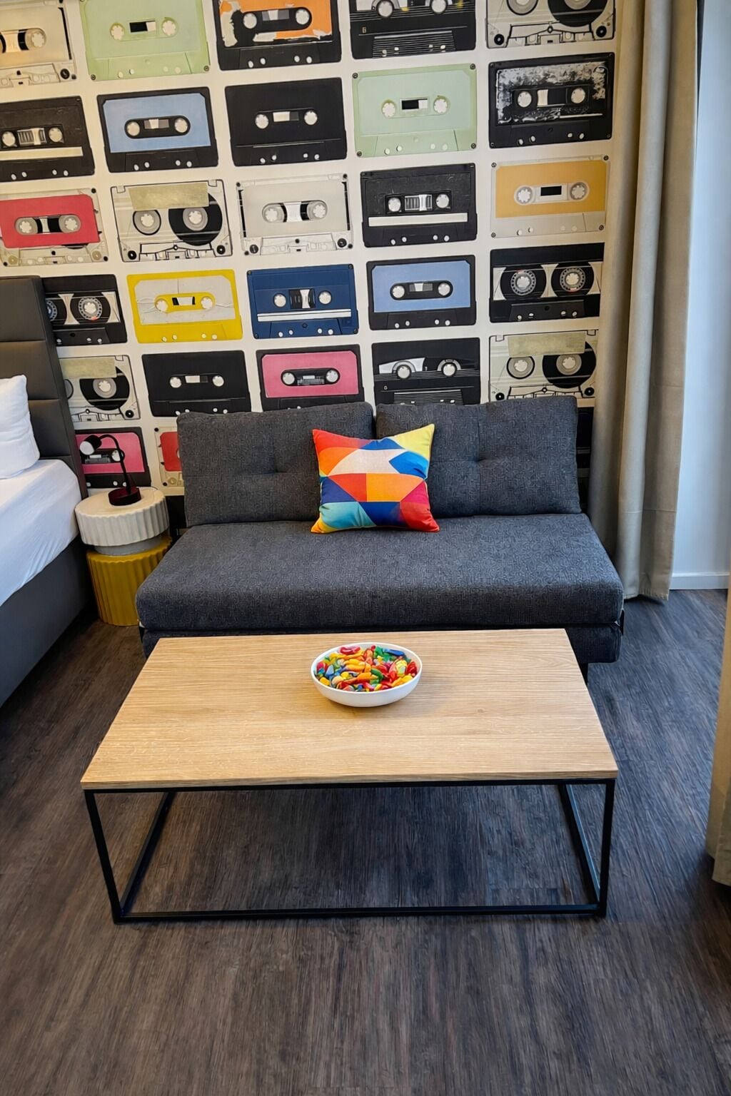 A modern room with a grey sofa, colorful cushions, a wooden coffee table with a bowl of fruit, and a wall decorated with vintage cassette tapes.