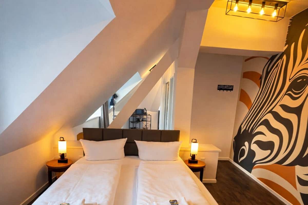 A hotel room with a large bed, two bedside tables, a zebra mural, and a sloped ceiling.