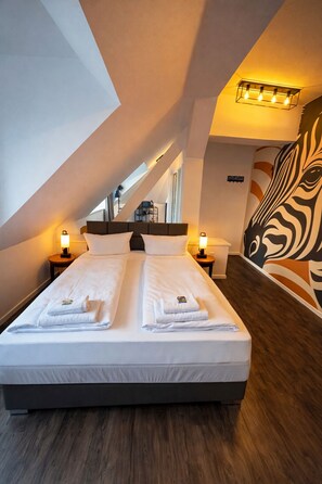 Una habitación de hotel con una cama grande, dos mesitas de noche, un mural de cebra y un techo inclinado.