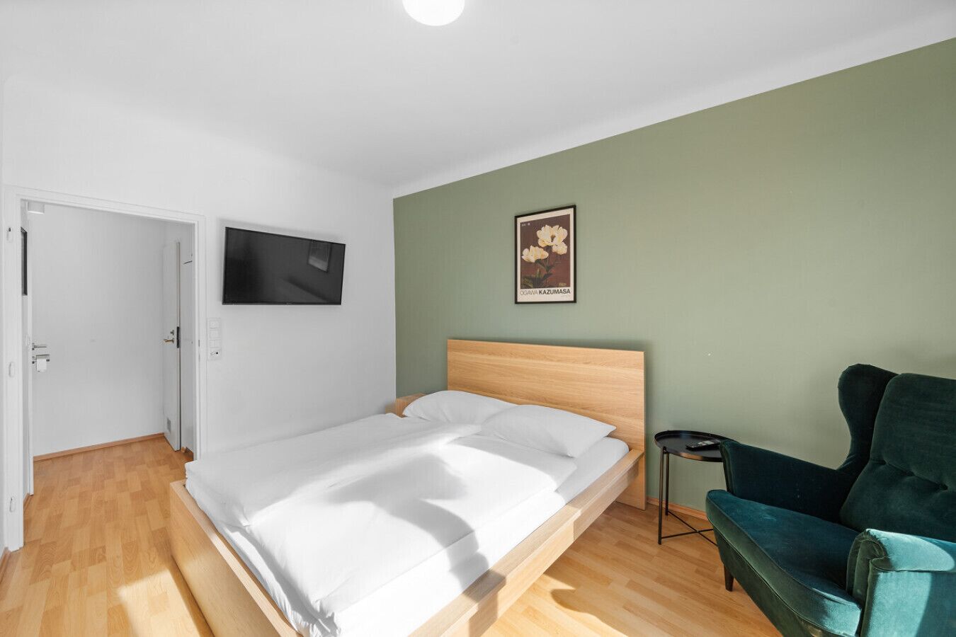 Un dormitorio con una cama de madera, una pared verde, un cuadro enmarcado y una silla color turquesa.